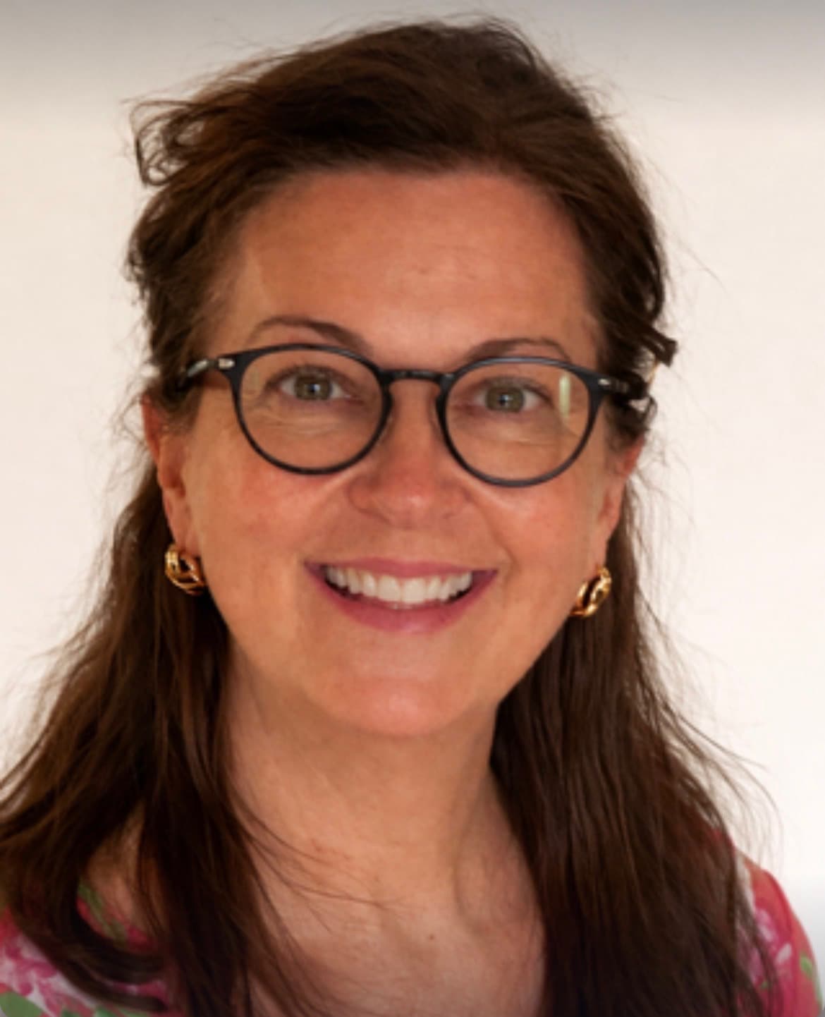 Smiling woman with brown hair, black-rimmed glasses, and gold earrings against a white background.
